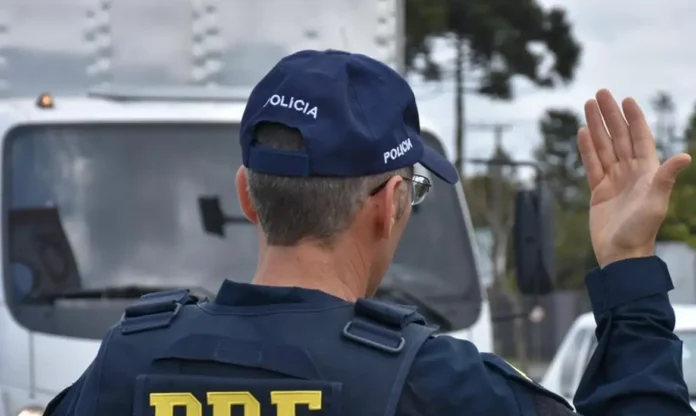Agente da Polícia Rodoviária Federal de costas, a sinalizar com a mão para orientar o trânsito rodoviário.