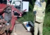 Bombeiros trabalham à noite ao lado de um carro vermelho totalmente destruído após um acidente. No chão está uma maca de transporte para a vítima.