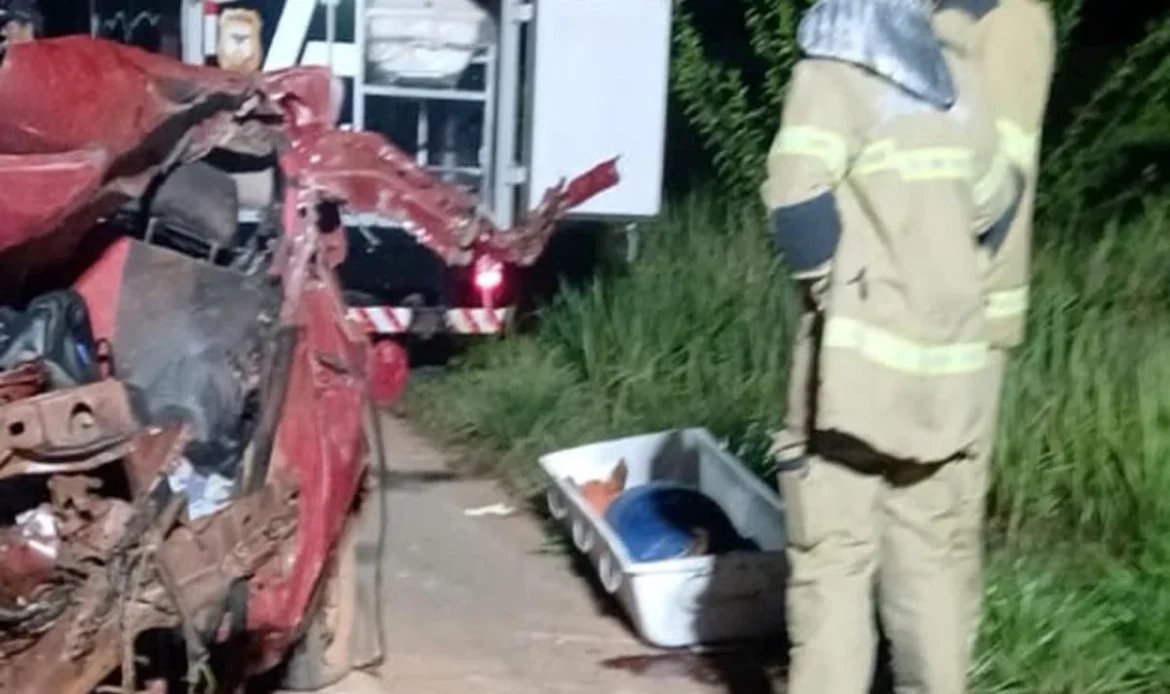Bombeiros trabalham à noite ao lado de um carro vermelho totalmente destruído após um acidente. No chão está uma maca de transporte para a vítima.