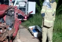 Motorista morre após bater de frente com ônibus na BR-319 em Porto Velho Bombeiros trabalham à noite ao lado de um carro vermelho totalmente destruído após um acidente. No chão está uma maca de transporte para a vítima.