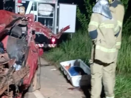 Motorista morre após bater de frente com ônibus na BR-319 em Porto Velho Bombeiros trabalham à noite ao lado de um carro vermelho totalmente destruído após um acidente. No chão está uma maca de transporte para a vítima.