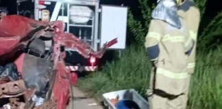 Motorista morre após bater de frente com ônibus na BR-319 em Porto Velho Bombeiros trabalham à noite ao lado de um carro vermelho totalmente destruído após um acidente. No chão está uma maca de transporte para a vítima.
