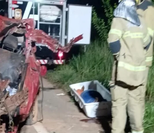 Bombeiros trabalham à noite ao lado de um carro vermelho totalmente destruído após um acidente. No chão está uma maca de transporte para a vítima.