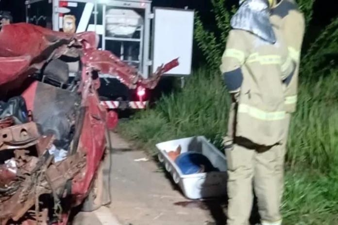 Bombeiros trabalham à noite ao lado de um carro vermelho totalmente destruído após um acidente. No chão está uma maca de transporte para a vítima.