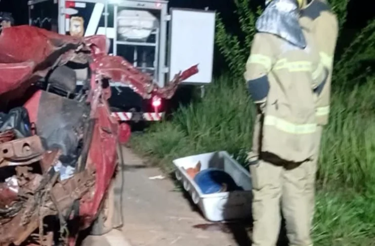 Motorista morre após bater de frente com ônibus na BR-319 em Porto Velho Bombeiros trabalham à noite ao lado de um carro vermelho totalmente destruído após um acidente. No chão está uma maca de transporte para a vítima.