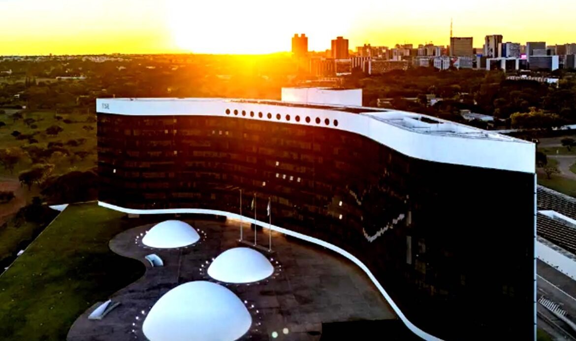 Vista aérea do edifício do Tribunal Superior Eleitoral (TSE) em Brasília, exibindo a sua arquitetura com fachada em curva negra e branca sob a luz de um pôr do sol.