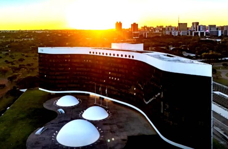 Vista aérea do edifício do Tribunal Superior Eleitoral (TSE) em Brasília, exibindo a sua arquitetura com fachada em curva negra e branca sob a luz de um pôr do sol.