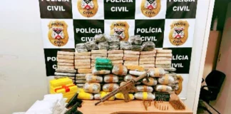 Mesa exibindo diversos pacotes de drogas empilhados, ladeados por seis revólveres, pistolas, um fuzil e dezenas de munições, com um painel da Polícia Civil ao fundo.