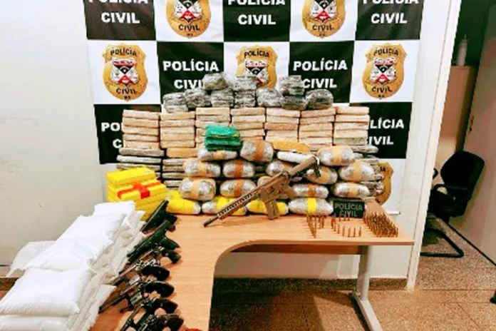 Fuzil, armas e pacotes de drogas apreendidos pela polícia Mesa exibindo diversos pacotes de drogas empilhados, ladeados por seis revólveres, pistolas, um fuzil e dezenas de munições, com um painel da Polícia Civil ao fundo.