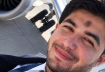 Selfie de um homem jovem com barba, vestindo uniforme de piloto com gravata, sorrindo levemente. Ao fundo, é possível ver parte do motor e do trem de pouso de um avião na pista.