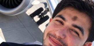 Selfie de um homem jovem com barba, vestindo uniforme de piloto com gravata, sorrindo levemente. Ao fundo, é possível ver parte do motor e do trem de pouso de um avião na pista.