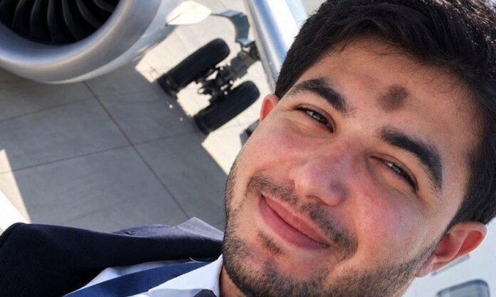 Selfie de um homem jovem com barba, vestindo uniforme de piloto com gravata, sorrindo levemente. Ao fundo, é possível ver parte do motor e do trem de pouso de um avião na pista.