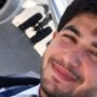 Selfie de um homem jovem com barba, vestindo uniforme de piloto com gravata, sorrindo levemente. Ao fundo, é possível ver parte do motor e do trem de pouso de um avião na pista.