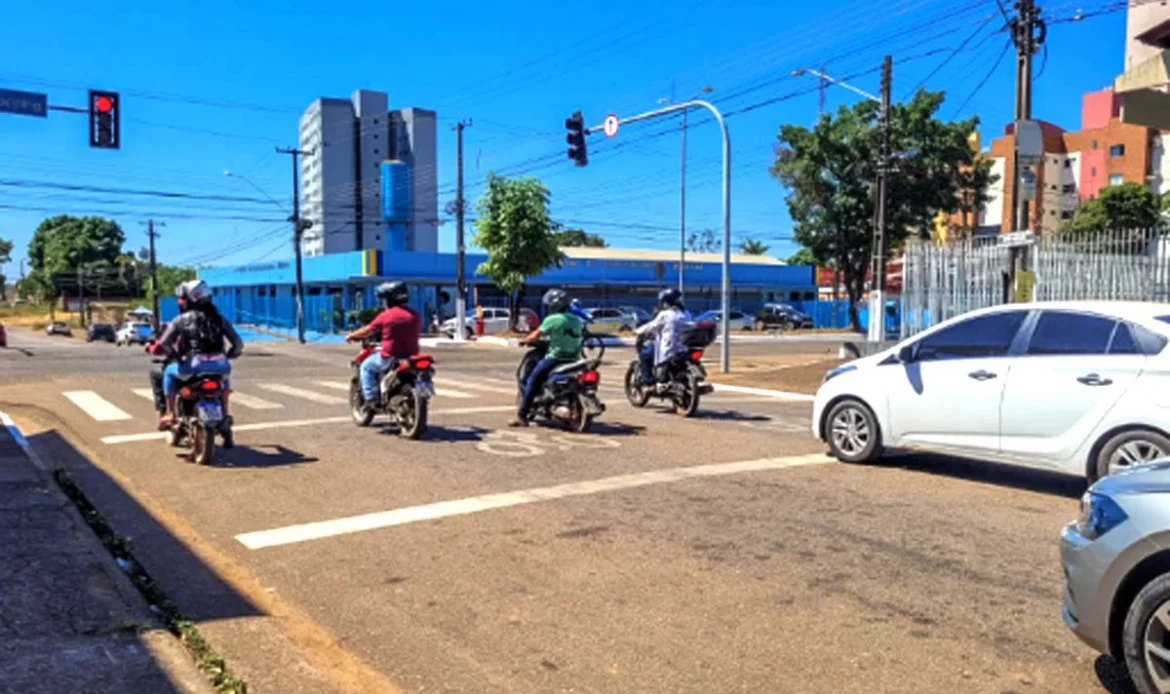 Quatro motociclistas aguardando o sinal abrir no semáforo em um cruzamento ensolarado, ao lado de carros brancos e pratas em uma avenida.