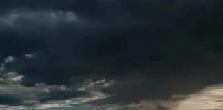 Céu escuro coberto por nuvens pesadas de tempestade sobre uma paisagem rural com silhuetas de árvores no horizonte.