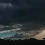 Céu escuro coberto por nuvens pesadas de tempestade sobre uma paisagem rural com silhuetas de árvores no horizonte.