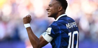 O jogador Mbappé sorrindo e comemorando em campo, vestindo a camisa azul da seleção francesa de futebol.