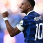O jogador Mbappé sorrindo e comemorando em campo, vestindo a camisa azul da seleção francesa de futebol.