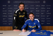 O jogador Cole Palmer assina um contrato sentado à mesa, com uma camisa do Chelsea ao lado, enquanto o técnico Xabi Alonso posa em pé, logo atrás dele.