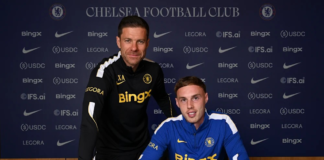 Cole Palmer renova com o Chelsea até 2034 O jogador Cole Palmer assina um contrato sentado à mesa, com uma camisa do Chelsea ao lado, enquanto o técnico Xabi Alonso posa em pé, logo atrás dele.