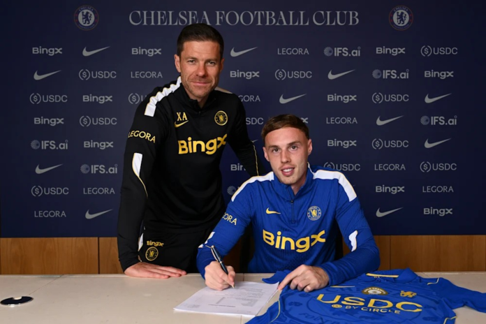 Cole Palmer e Xabi Alonso na assinatura da renovação O jogador Cole Palmer assina um contrato sentado à mesa, com uma camisa do Chelsea ao lado, enquanto o técnico Xabi Alonso posa em pé, logo atrás dele.