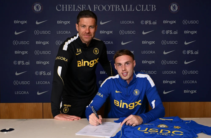 O jogador Cole Palmer assina um contrato sentado à mesa, com uma camisa do Chelsea ao lado, enquanto o técnico Xabi Alonso posa em pé, logo atrás dele.