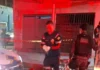 Profissional do Samu de luvas brancas e policial militar do batalhão de Choque trabalhando em uma cena de crime à noite, com área isolada por fita zebrada.