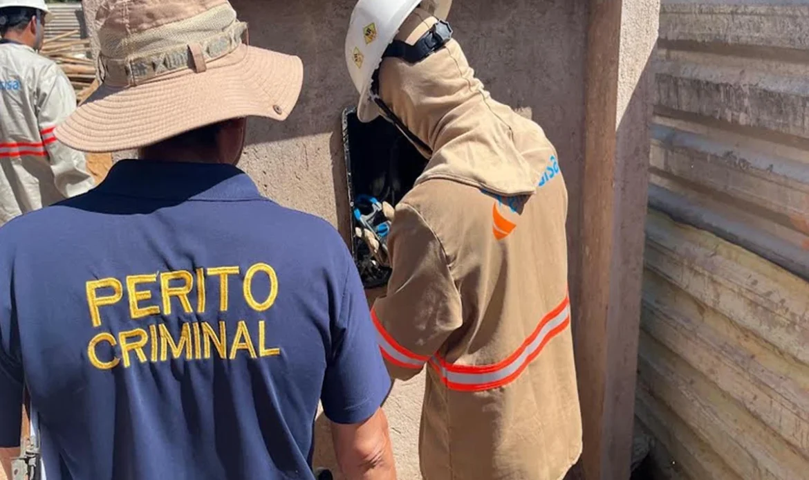 Um perito criminal vestindo uma camisa azul escura com o texto "PERITO CRIMINAL" em amarelo acompanha um técnico da Energisa que inspeciona uma caixa de energia.