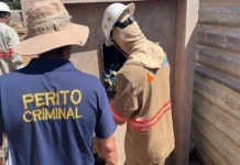 Um perito criminal vestindo uma camisa azul escura com o texto "PERITO CRIMINAL" em amarelo acompanha um técnico da Energisa que inspeciona uma caixa de energia.