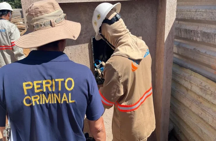 Furto de energia causa rombo de R$ 200 milhões nos cofres públicos de Rondônia Um perito criminal vestindo uma camisa azul escura com o texto "PERITO CRIMINAL" em amarelo acompanha um técnico da Energisa que inspeciona uma caixa de energia.