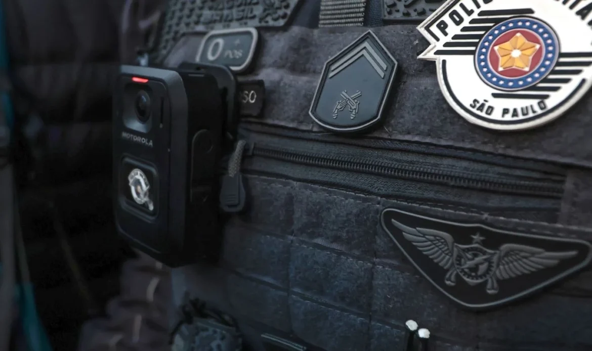 Colete tático escuro de um agente da Polícia Militar de São Paulo, equipado com uma câmara corporal (bodycam) e ostentando as insígnias oficiais da corporação.