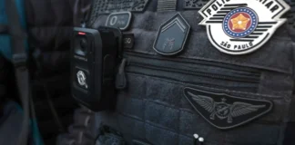 Colete tático escuro de um agente da Polícia Militar de São Paulo, equipado com uma câmara corporal (bodycam) e ostentando as insígnias oficiais da corporação.