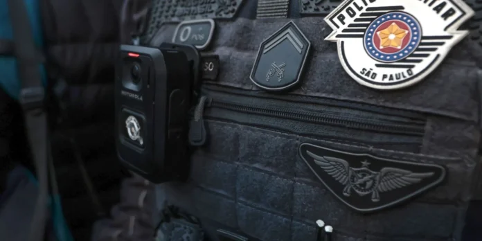 Agente da Policia Militar de Sao Paulo com bodycam Colete tático escuro de um agente da Polícia Militar de São Paulo, equipado com uma câmara corporal (bodycam) e ostentando as insígnias oficiais da corporação.