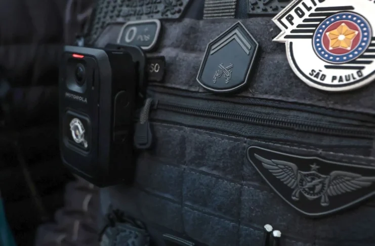 Colete tático escuro de um agente da Polícia Militar de São Paulo, equipado com uma câmara corporal (bodycam) e ostentando as insígnias oficiais da corporação.