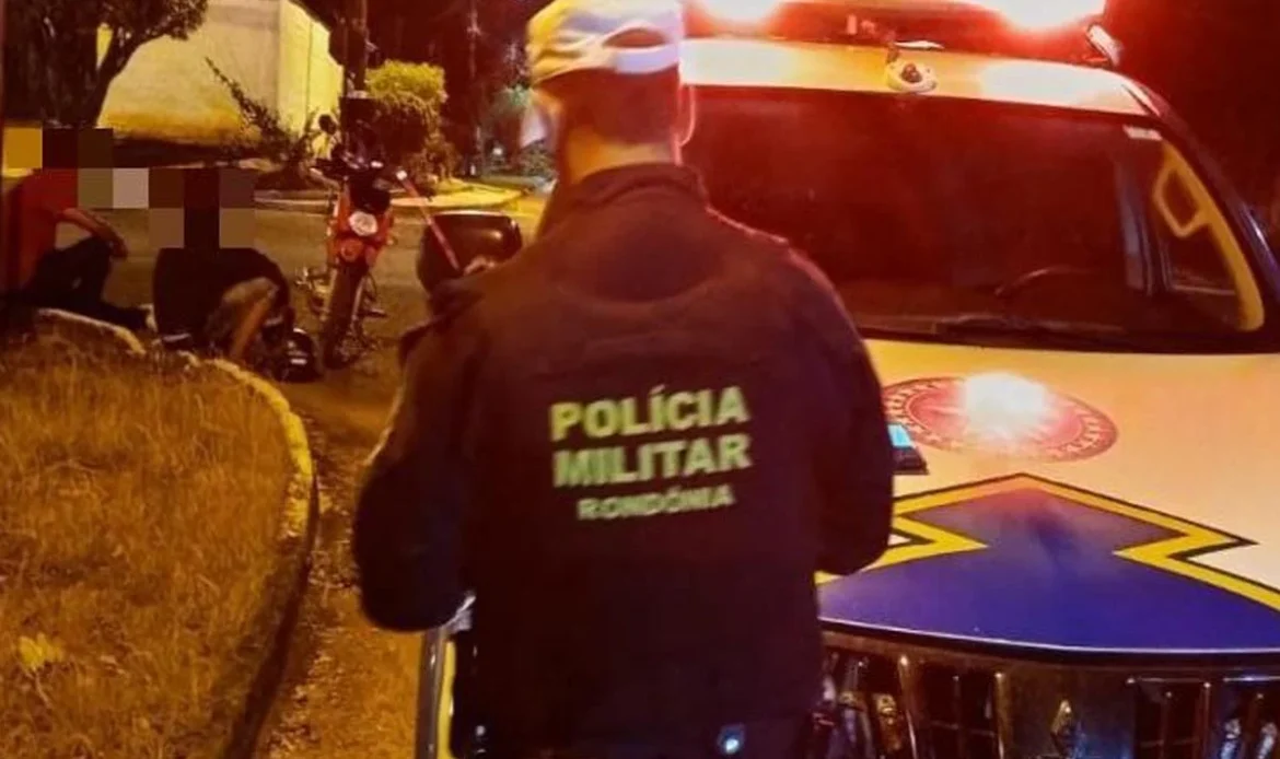 Policial militar de Rondônia de costas, ao lado de uma viatura com as luzes ligadas à noite, olhando para uma moto caída e pessoas sentadas na calçada.