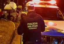 Policial militar de Rondônia de costas, ao lado de uma viatura com as luzes ligadas à noite, olhando para uma moto caída e pessoas sentadas na calçada.
