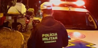 Policial militar de Rondônia de costas, ao lado de uma viatura com as luzes ligadas à noite, olhando para uma moto caída e pessoas sentadas na calçada.
