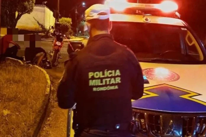 Policial militar de Rondônia de costas, ao lado de uma viatura com as luzes ligadas à noite, olhando para uma moto caída e pessoas sentadas na calçada.