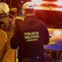 Policial militar de Rondônia de costas, ao lado de uma viatura com as luzes ligadas à noite, olhando para uma moto caída e pessoas sentadas na calçada.