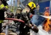 Piloto morre em queda de avião no pátio do metrô de BH Bombeiro militar, utilizando capacete amarelo e equipamento de proteção respiratória, combate as chamas com uma mangueira de água sobre os destroços de uma aeronave acidentada.
