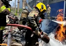 Bombeiro militar, utilizando capacete amarelo e equipamento de proteção respiratória, combate as chamas com uma mangueira de água sobre os destroços de uma aeronave acidentada.