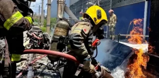 Bombeiro militar, utilizando capacete amarelo e equipamento de proteção respiratória, combate as chamas com uma mangueira de água sobre os destroços de uma aeronave acidentada.