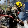 Bombeiro militar, utilizando capacete amarelo e equipamento de proteção respiratória, combate as chamas com uma mangueira de água sobre os destroços de uma aeronave acidentada.