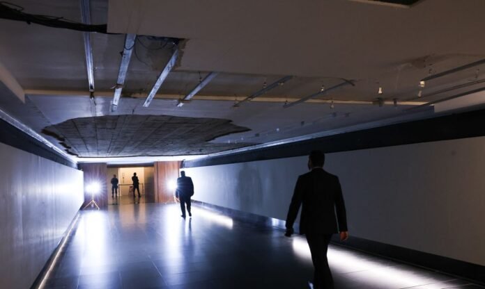 Teto do Tunel do Tempo desaba na Camara dos Deputados Corredor do Túnel do Tempo na Câmara dos Deputados com um buraco visível no teto devido à queda do reboco, iluminado por holofotes e com a silhueta de homens a caminhar.