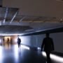 Corredor do Túnel do Tempo na Câmara dos Deputados com um buraco visível no teto devido à queda do reboco, iluminado por holofotes e com a silhueta de homens a caminhar.