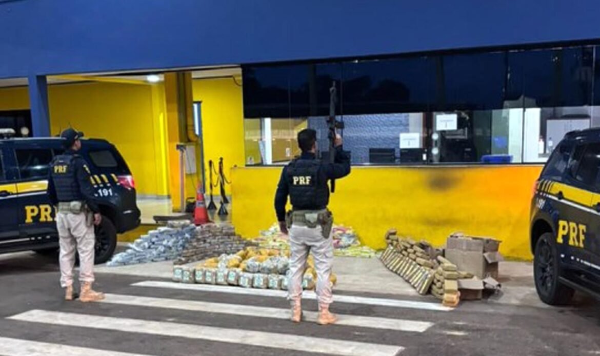 Agentes da Polícia Rodoviária Federal armados em frente ao posto policial, junto a duas viaturas pretas e dezenas de pacotes de droga apreendidos dispostos no chão.