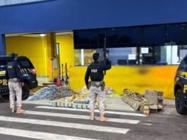 Agentes da Polícia Rodoviária Federal armados em frente ao posto policial, junto a duas viaturas pretas e dezenas de pacotes de droga apreendidos dispostos no chão.