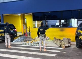 Agentes da Polícia Rodoviária Federal armados em frente ao posto policial, junto a duas viaturas pretas e dezenas de pacotes de droga apreendidos dispostos no chão.