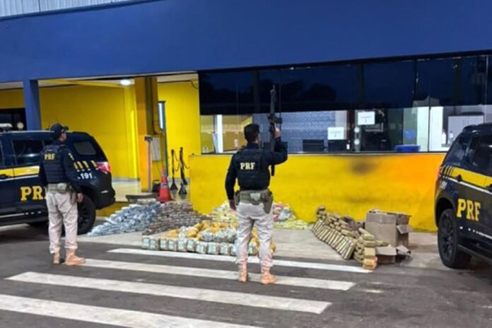 PRF apreende quase 500 kg de drogas em Pimenta Bueno Agentes da Polícia Rodoviária Federal armados em frente ao posto policial, junto a duas viaturas pretas e dezenas de pacotes de droga apreendidos dispostos no chão.