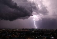 Trovoadas ganham força e atingem Rondônia e mais quatro estados do Norte Céu noturno coberto por nuvens escuras de tempestade sobre uma cidade, com um raio forte caindo e iluminando o horizonte.